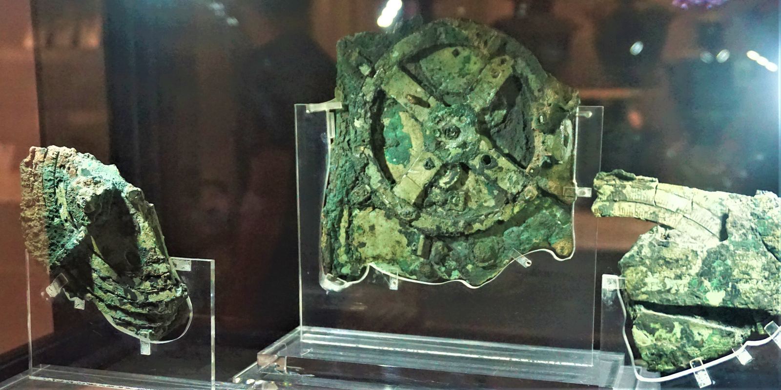 Green corroded bronze fragments of the Antikythera mechanism mounted on clear stands in a glass case at the National Archaeological Museum in Athens