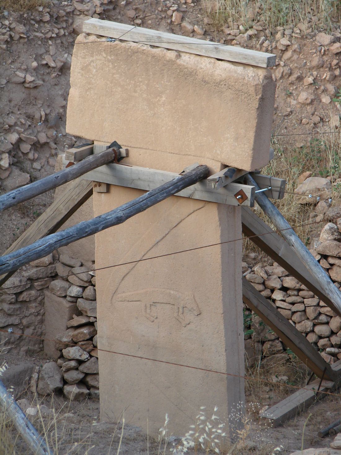 A tall T-shaped limestone pillar at Göbekli Tepe with a carved arm running down its side and a fox relief, supported by wooden props