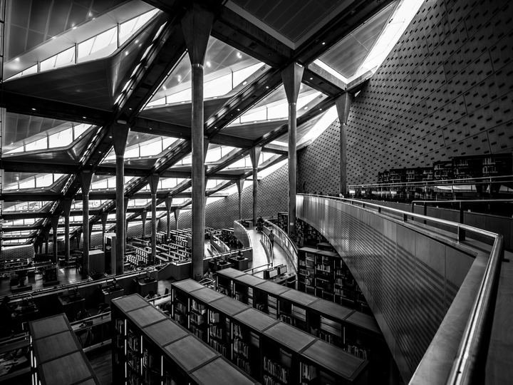 Black and white view of the vast terraced reading room of the Bibliotheca Alexandrina, with rows of desks under a slanted roof of skylights held up by tall slender columns