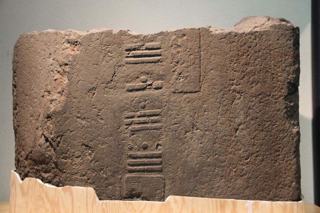A weathered stone stela fragment from Tres Zapotes with a vertical column of carved bar-and-dot numerals, displayed in a museum