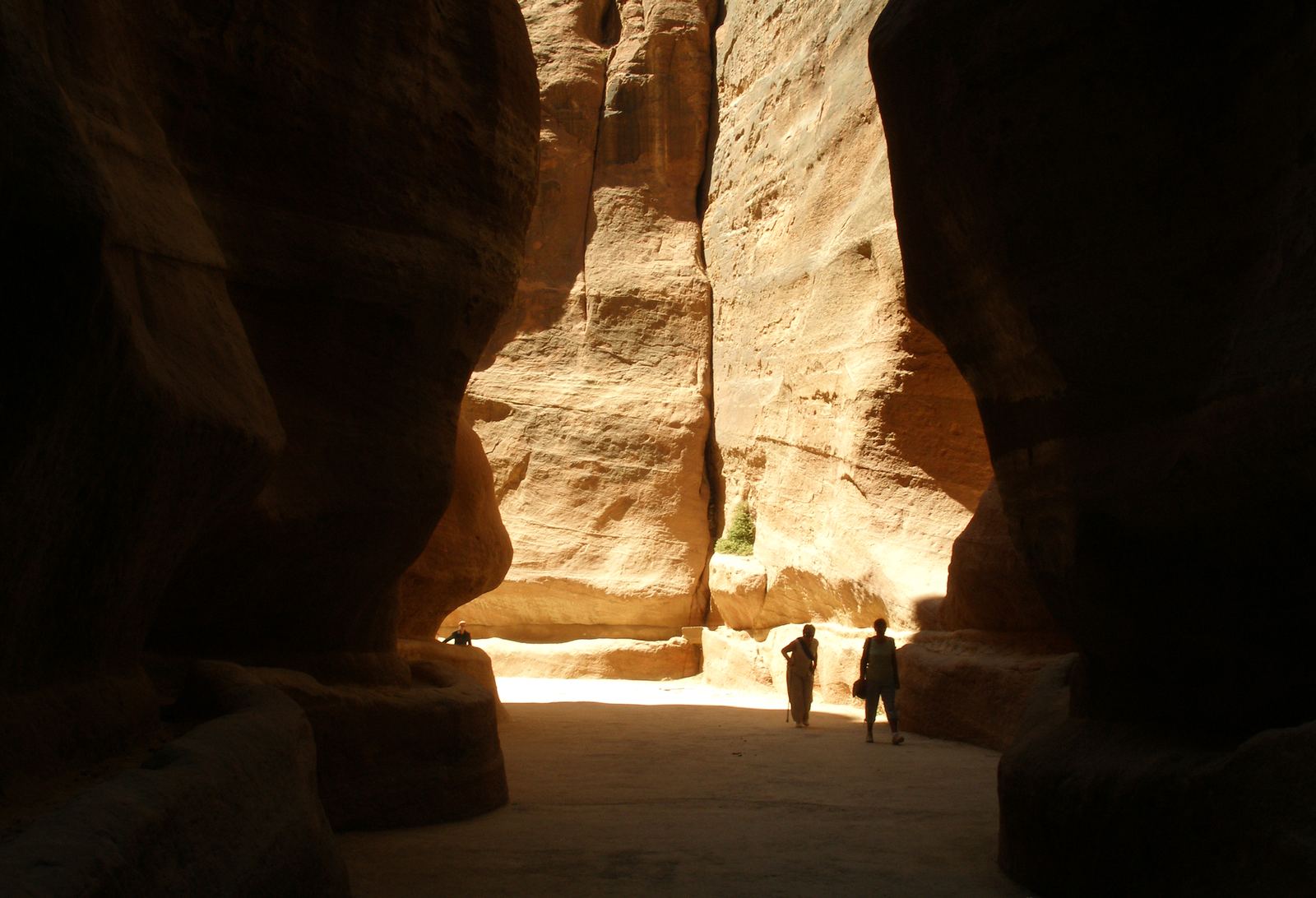 The narrow Siq canyon at Petra, Jordan, with towering curved walls of pink and orange sandstone and two small figures walking toward a band of light