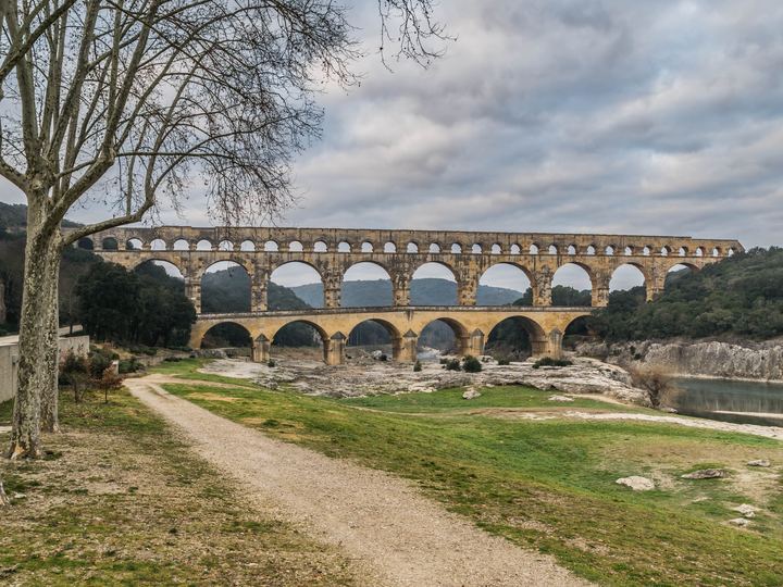 The three tiers of honey-colored stone arches of the Pont du Gard crossing the Gardon river valley under a cloudy sky, with a gravel path in the foreground