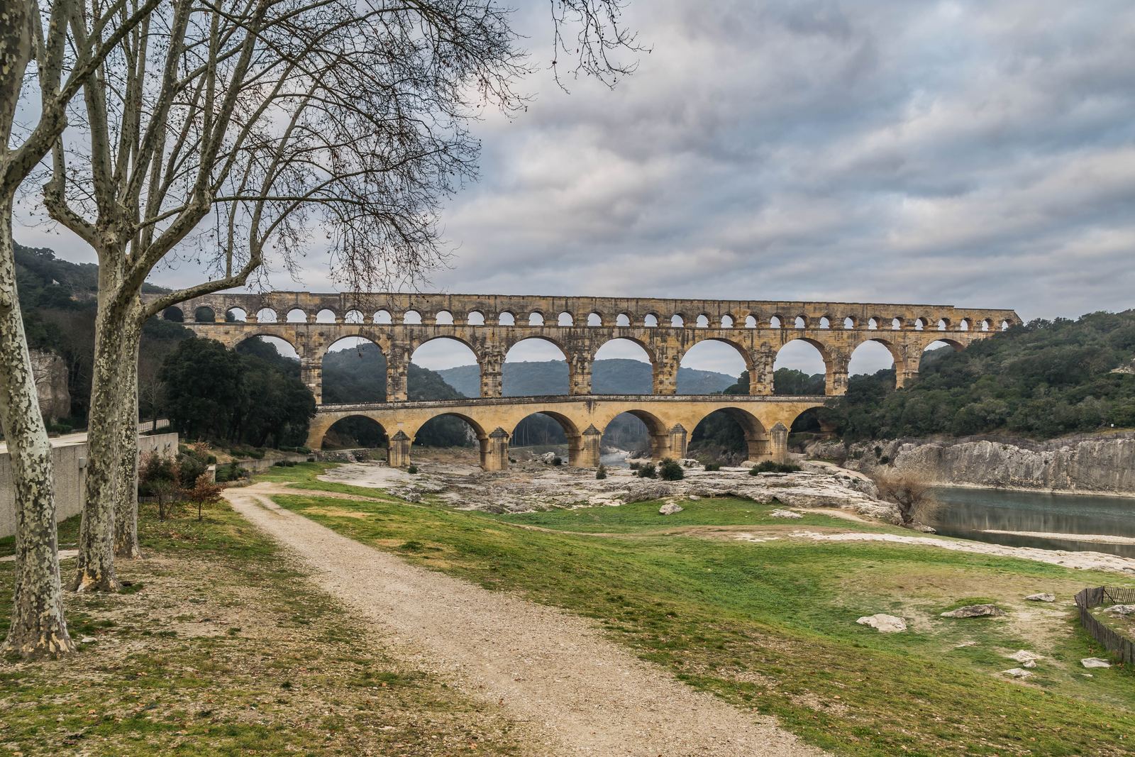 The three tiers of honey-colored stone arches of the Pont du Gard crossing the Gardon river valley under a cloudy sky, with a gravel path in the foreground